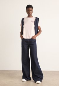 T-shirt en coton rose avec un petit motif, un sweat sans manches bleu marine noué sur les épaules, et un pantalon en denim large bleu foncé. Baskets blanches.