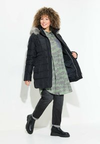 Abrigo acolchado negro con capucha con borde de piel, sobre una camisa verde con estampado. Combinado con jeans oscuros y botas de plataforma negras. Ajuste casual, cierre con cremallera.