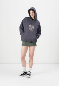 Mørkegrå hettegenser med stor frontgrafikk, kombinert med et grønt miniskjørt og svarte sneakers. Hvite ankelstrømper fullfører antrekket.