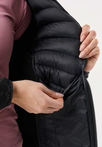 Personne portant une chemise manches longues mauve tenant ouverte une veste matelassée noire, montrant une poche intérieure avec un rabat.
