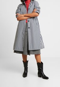 Manteau long à motif vichy bleu et blanc avec manches trois-quarts, jupe noire à carreaux, et bottes de cow-boy noires.