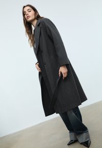 Mörkgrå ullrock med dubbelbröstdesign, breda slag och sidofickor, tillsammans med mörka denimjeans och spetsiga svarta klackar.