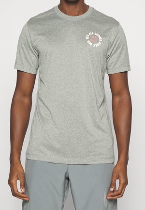 Mann trägt ein graues T-Shirt mit kurzen Ärmeln und einem kleinen runden Logo auf der Brust mit der Aufschrift "IT'S ALL GONNA WORK OUT", das ein rotes Smiley-Gesicht umgibt.