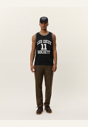 Black mesh tank top with white print "LES DEUX 11 SOCIETY," paired with brown plaid trousers and dark shoes, worn with a matching cap.