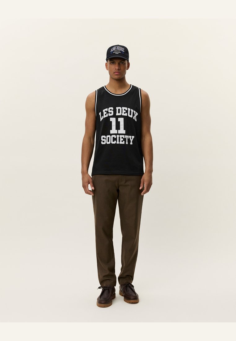 Black mesh tank top with white print "LES DEUX 11 SOCIETY," paired with brown plaid trousers and dark shoes, worn with a matching cap.