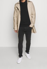 Béžový trenčkot přes černou mikinu s kapucí, v kombinaci s černými džínami a bílými teniskami. Má knoflíky a volný střih.