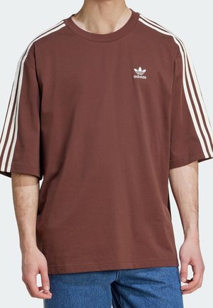 Mann trägt braunes Adidas-T-Shirt mit weißen drei Streifen an den Ärmeln und Logo auf der Brust, kombiniert mit blauen Jeans.