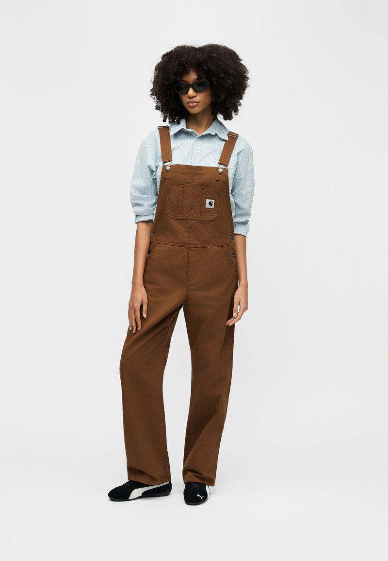 Woman with curly hair wearing black sunglasses, light blue shirt, brown overalls, and black Puma sneakers standing against a plain background.