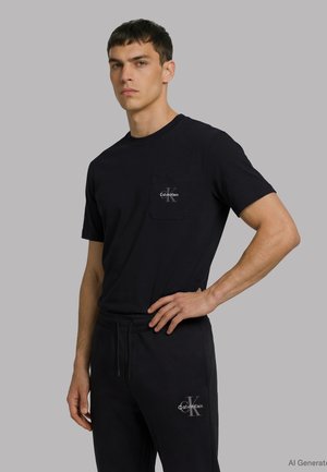 Mężczyzna noszący czarną koszulkę i spodnie Calvin Klein z logo, stojący z jedną ręką na biodrze na jednolitym szarym tle.