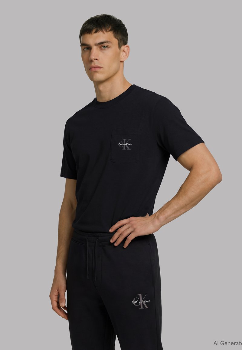 Άνδρας που φοράει μαύρο t-shirt και παντελόνι Calvin Klein με λογότυπα, στέκεται με το ένα χέρι στη μέση σε απλό γκρι φόντο.