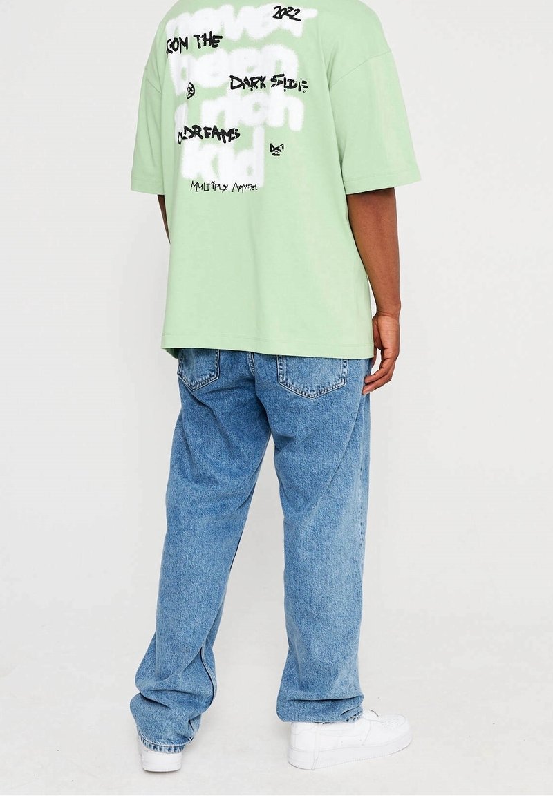 T-shirt oversize vert clair avec des graphismes de texte noir et blanc au dos, associé à un jean denim bleu ample et des baskets blanches.
