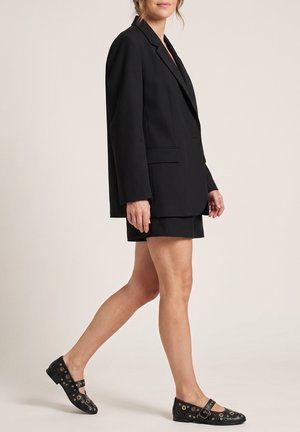 Kobieta w oversize'owym czarnym blezerze i dopasowanej mini spódnicy, idzie w czarnych balerinkach Mary Jane z metalowymi oczkami.