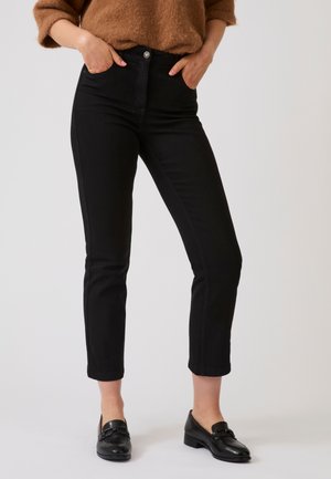 Donna che indossa pantaloni neri slim fit alla caviglia abbinati a mocassini neri e a un maglione marrone, con le mani nelle tasche, in piedi su sfondo bianco.