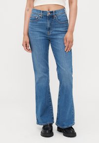 Modre flared jeans iz deprine z visokim pasom, z sprednjimi žepi, bronastimi dodatki in nekoliko zbledelim videzom. Obuti v črne čevlje.