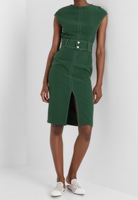 Robe verte ajustée avec des manches courtes, une fente à l'avant et une taille ceinturée. Présente des coutures contrastées et une texture lisse.