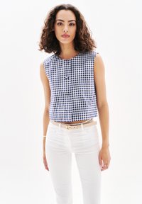 Top senza maniche a scacchi blu e bianchi con scollatura rotonda e bottoni frontali, abbinato a jeans bianchi aderenti e una cintura beige.