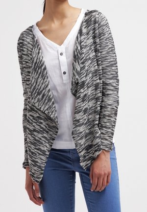 Kobieta ubrana w czarno-biały kardigan z otwartym przodem, nałożony na białą koszulę z guzikami i niebieskie dżinsy, stoi na neutralnym tle.