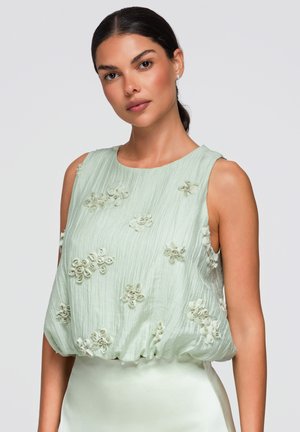 Donna con capelli scuri raccolti indossa un top verde chiaro senza maniche con applicazioni floreali testurizzate su uno sfondo uniforme.
