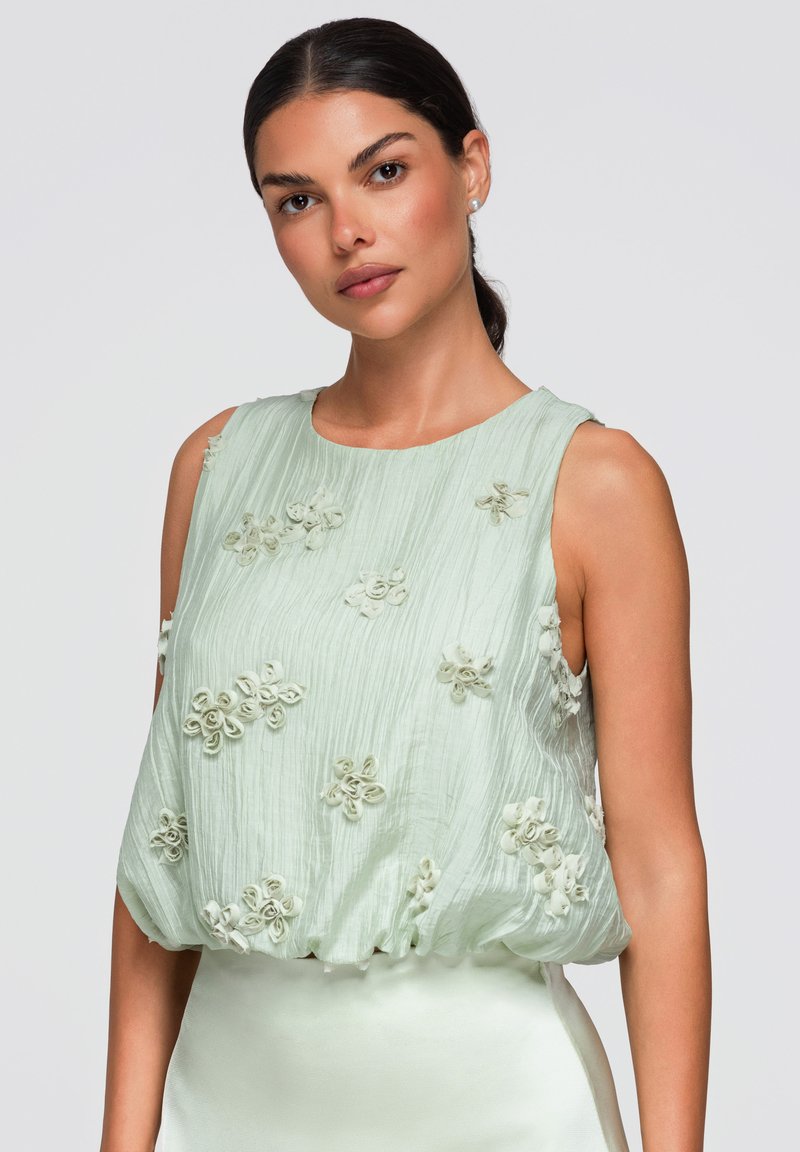 Femme aux cheveux foncés tirés en arrière, portant un haut sans manches vert clair avec des applications florales texturées sur un fond uni.