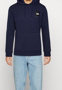 Navy modrá mikina s kapucí z měkkého materiálu, s přední kapsou ve stylu klokan, kapucí se stahovací šňůrkou a nášivkou logo na hrudi.