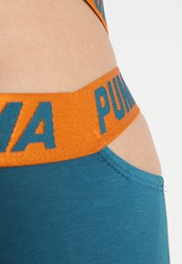 Tyrkysové šortky z látky s širokým oranžovým elastickým pasem, na kterém je v tyrkysovém písmu napsáno "PUMA". Hladký povrch a přiléhavý design.