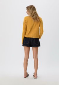 Frau, die mit dem Rücken zur Kamera steht, trägt einen senfgelben Pullover, dunkle Jeansshorts und gemusterte Ballerinas vor einem schlichten Hintergrund.
