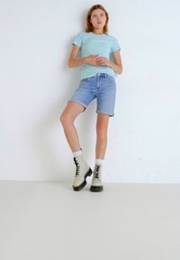 Šviesiai mėlynas prigludęs marškinėliai, šviesiai džinsiniai šortai, ir kreminės spalvos raištiniai kulkšninių batai su juodais padais. Modelis stovi prieš baltą foną.