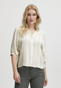 Blusa abotonada en color crema claro con rayas verticales, hecha de una tela suave. Presenta un escote en V y mangas abullonadas de tres cuartos.