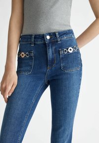 Hoge taille blauwe denim jeans met een soepele textuur, voorzien van twee decoratieve metalen accenten op de achterzakken en klassieke stiksels.
