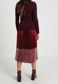 Bordeaux kleurige, getailleerde sweater gecombineerd met een geplooide, twee-toon midi rok. De rok heeft glanzende, diep rode en zachte roze secties, met textuurcontrast.