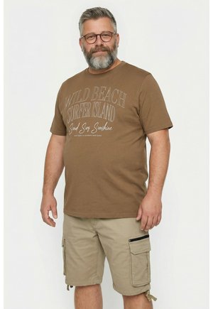 Mężczyzna z brodą i okularami, ubrany w brązowy T-shirt „Wild Beach Surfer Island” i beżowe szorty typu cargo, stoi na białym tle.