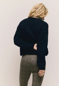 Teksturowany granatowy sweter z obniżonymi ramionami, zestawiony z brązowymi spodniami w kratę. Outfit charakteryzuje się luźnym krojem i neutralnymi odcieniami.