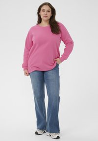 Femme debout avec une main dans la poche, portant un sweat-shirt rose, un jean bleu et des baskets blanches sur un fond blanc.