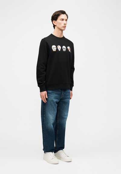 Άνδρας που φορά μαύρο φούτερ με τέσσερα πολύχρωμα σχέδια κρανίων στο στήθος, μπλε τζιν και λευκά αθλητικά, στέκεται μπροστά σε λευκό φόντο.