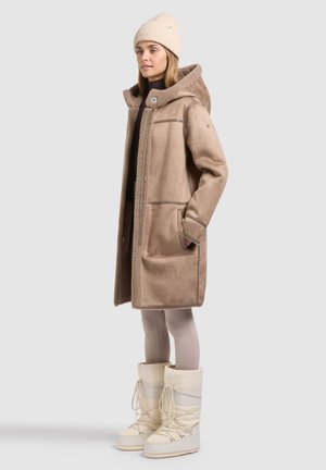 Femme portant un manteau en peau lainée beige à capuche, un bonnet tricoté crème, des leggings clairs et des bottes d’hiver blanches, debout de profil sur un fond neutre.