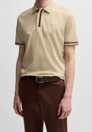 Personne portant un polo beige avec une bordure rayée marron et crème aux manches et au col, associé à un pantalon marron et une ceinture noire.