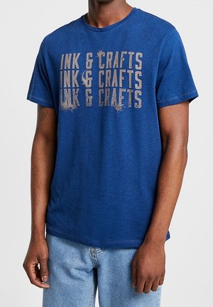 Hombre vistiendo una camiseta azul con texto beige desgastado "INK & CRAFTS" repetido tres veces, combinado con jeans de color azul claro.