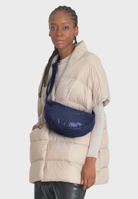 Béžová péřová vesta s krátkými rukávy, která má přes tělo přehozenou hluboce námořnickou tašku z hladkého, lesklého materiálu.