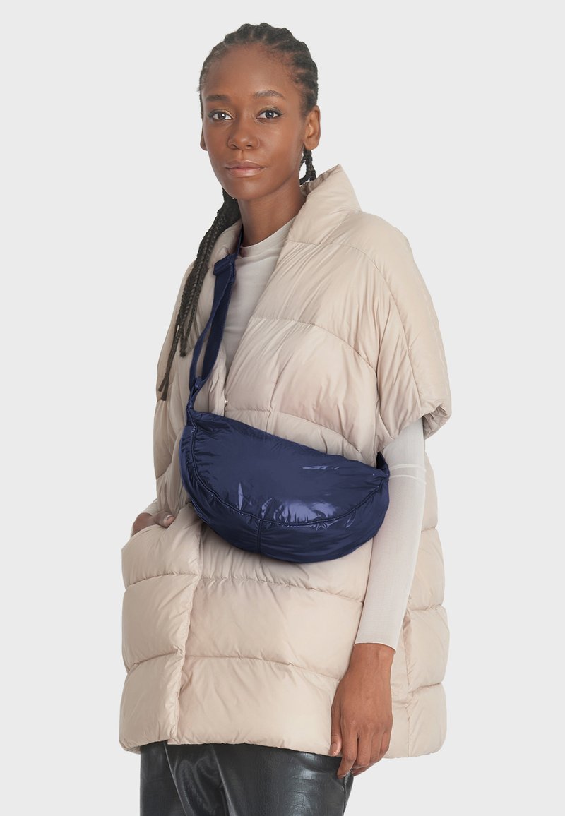 Béžová péřová vesta s krátkými rukávy, která má přes tělo přehozenou hluboce námořnickou tašku z hladkého, lesklého materiálu.