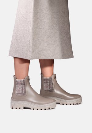 Personne portant des bottines de pluie beige à la cheville avec des semelles épaisses et des panneaux latéraux en tricot, associées à une jupe grise claire longueur genou.