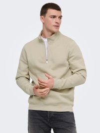 Homme portant un pull beige à fermeture éclair jusqu'au quart et un jean gris foncé, regardant sur le côté avec les mains jointes devant lui.