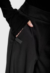 Czarne dopasowane spodnie z gładkiego materiału, z boczną kieszenią i małą gumowaną metką. Częściowo widoczny detal z rozszerzanym rękawem.