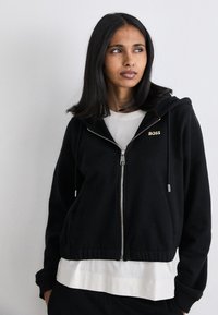 Crna zip-up dukserica s uskim rubom, s zlatnim "BOSS" logotipom, i s vezicama, izrađena od mekanog materijala. Nosiva preko bijele majice.