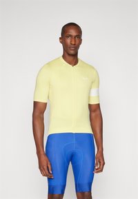Kurzarm-Radtrikot aus hellgelbem Stoff mit einem Frontreißverschluss und weißen Akzenten an den Ärmeln, kombiniert mit blauen gepolsterten Shorts.