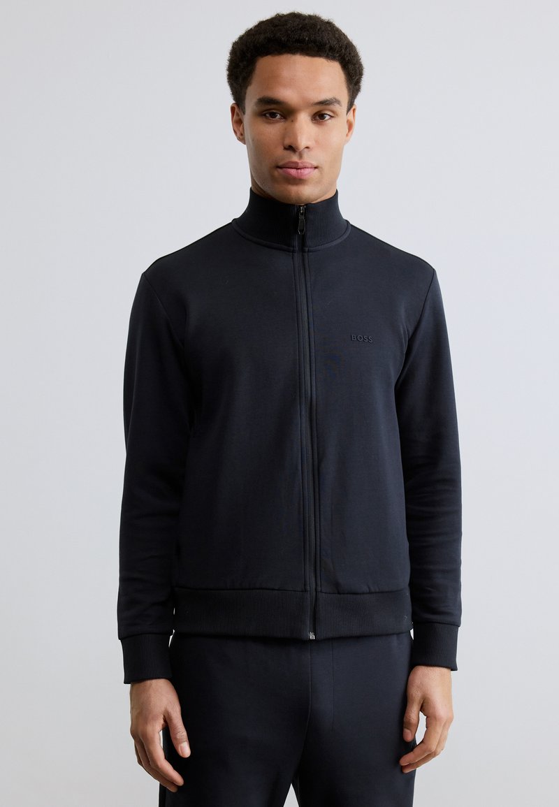 Άνδρας με μαύρο μπουφάν με φερμουάρ και ριμπ μανσέτες και γιακά, στέκεται μπροστά σε ένα απλό ανοιχτό γκρι φόντο.