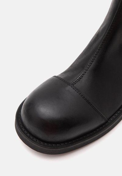 Gros plan sur une botte en cuir noir, montrant l’extrémité arrondie, le détail des coutures et la semelle noire robuste sur un fond clair.