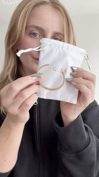 Bangle in oro esposto in un sacchetto bianco con cordoncino con il logo "GUESS". Il sacchetto ha una texture liscia, mentre il bracciale presenta una finitura lucida.
