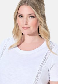 Bílý tričko s jemnými krajkovými detaily na ramenou, kulatým výstřihem, krátkými rukávy a hladkým, lehkým materiálem.