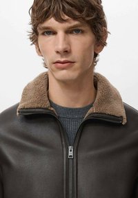 Giubbotto di pelle marrone con colletto in shearling beige, chiusura con zip sul davanti e maglione grigio testurizzato sotto, con un ritratto in primo piano.