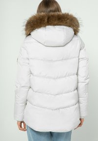 Bílá péřová bunda s kapucí lemovanou kožešinou, s prošívaným designem a žebrovanými akcenty, nošená přes světle modré džíny.